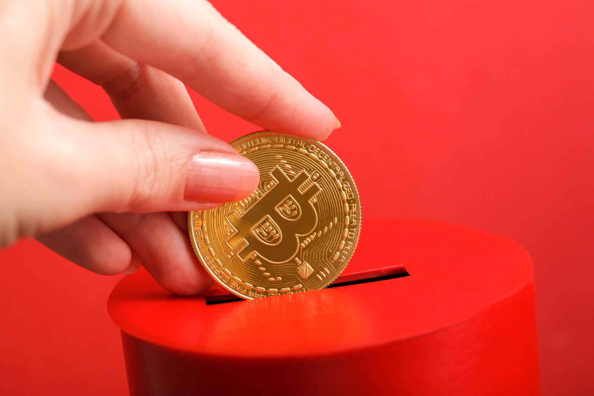 Female hand puts gold coin with bitcoin symbol into slot of red donation box. Concept of donorship, life saving or charity. Concept of sincere devotion to faith. Close-up shot