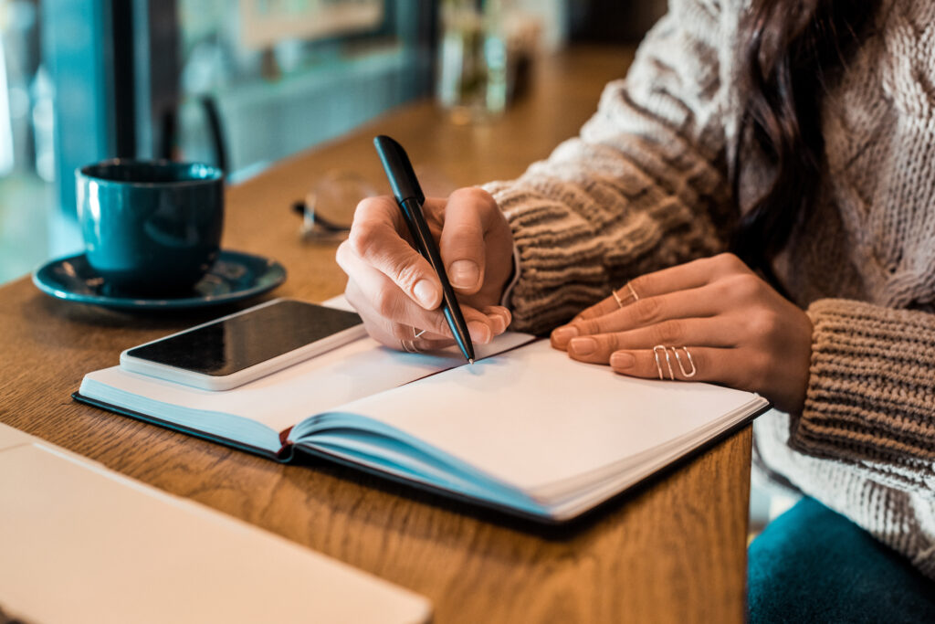 cropped view of freelancer writing in planner with smartphone in cafe