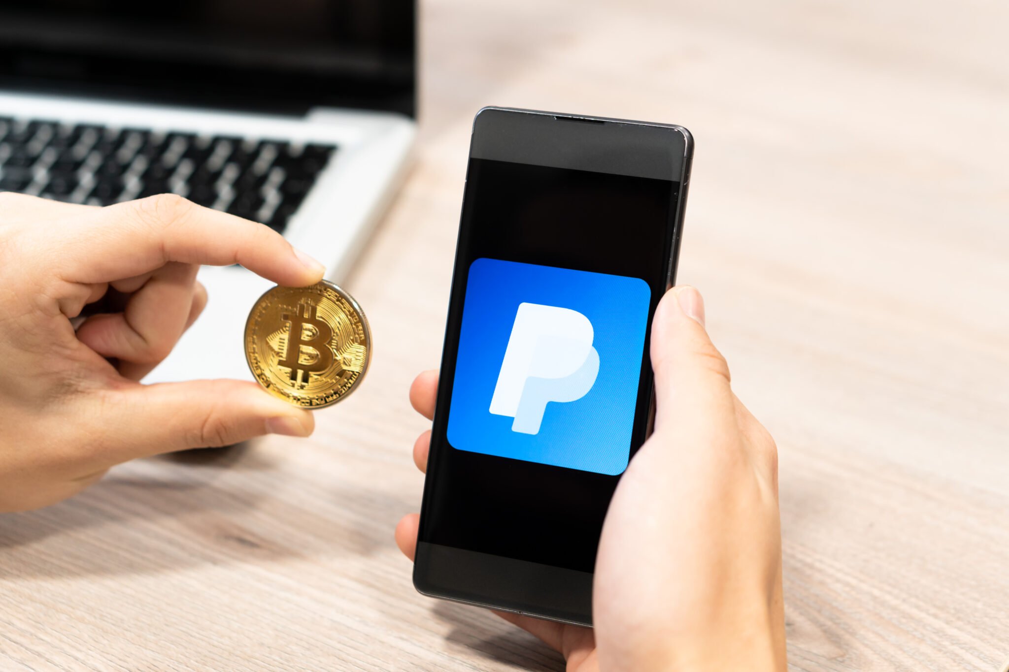Human hand holding mobile phone and Bitcoin coin - paypal logo displayed on phone,, computer laptop in the background. Slovenia 13.02.2019