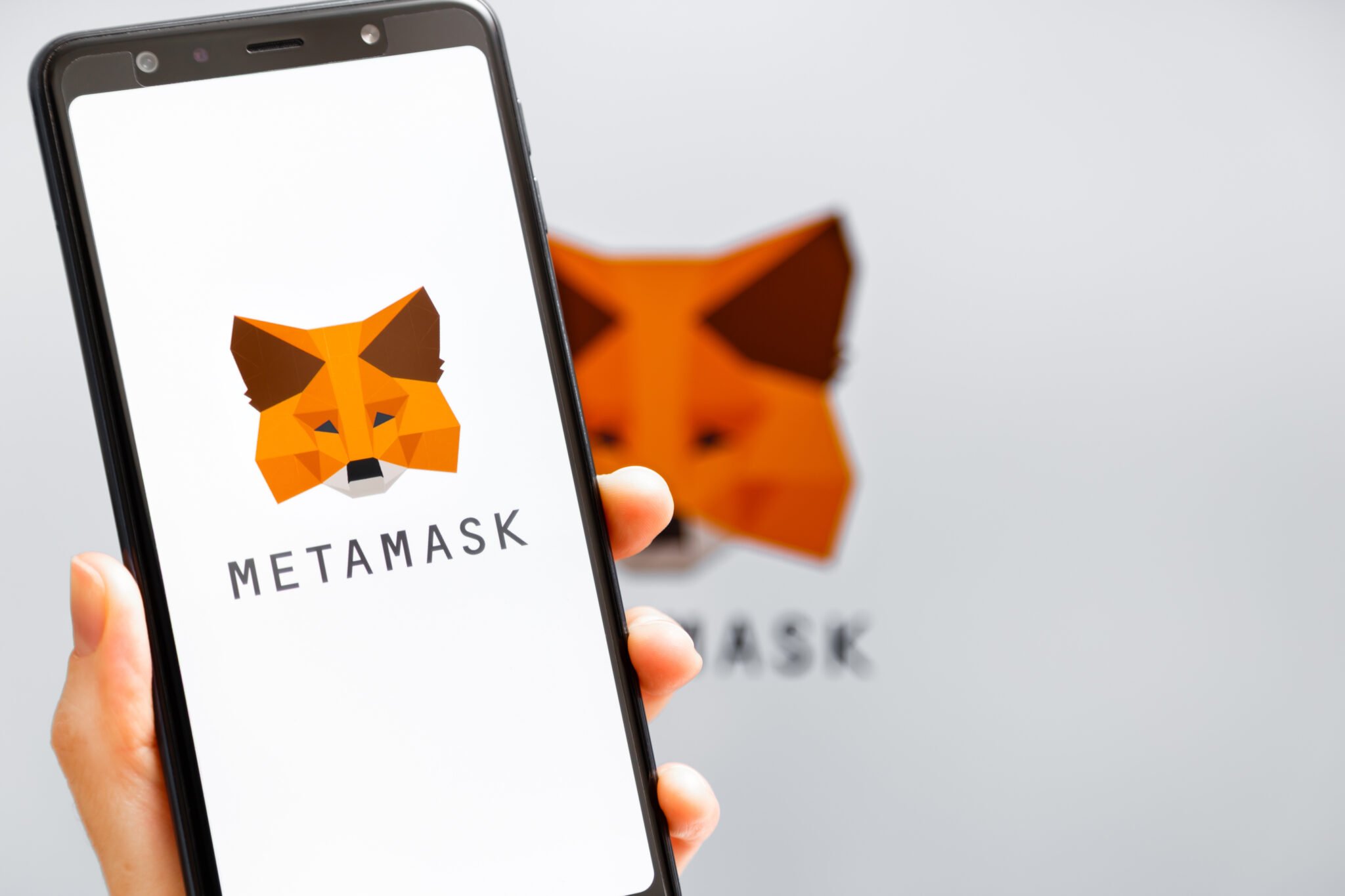 Ukraine, Odessa - October, 9 2021: Hand holding mobile with MetaMask app running at smartphone screen with MetaMask logo at background. MetaMask is software crypto wallet