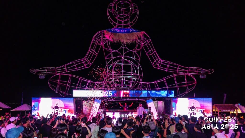 Nighttime concert and show at Coinfest Asia 2025 in Bali, featuring a glowing human-shaped statue and a lively crowd.